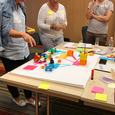 A colorful DIY session around the table with postits, markers, and prototyping materials. Three participants standing around the table brainstorming.