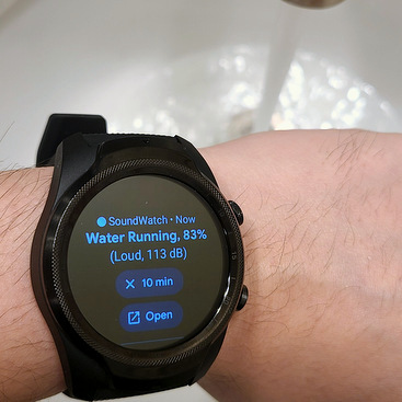 A user is wearing a smartwatch in front of water running down a sink. The smartwatch displays the identified sound as 'water running' with a classification confidence of 83%.