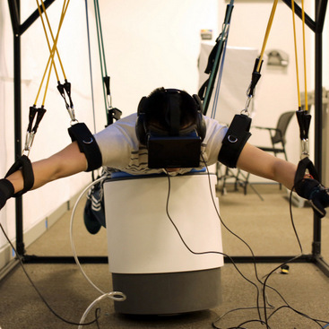 A person wearing an Oculus Rift VR headset and lying down on a soft inflatable mattress. His limbs are suspended through elastic bands in a rig to simulate swimming.