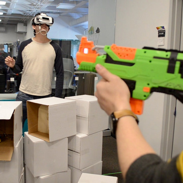 Two people are wearing an Oculus Rift VR headset. One person is defending an incoming virtual snowball launched by another person using a toy gun.